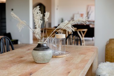 Light wood tabletop with a grey and black ceramic vase with stalks of wispy grass in it. Behind the vase, there's a glass pitcher and glasses.