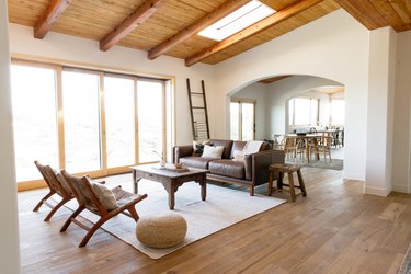 A large, open living room with white walls, sliding glass doors, and a high ceiling with wooden beams. There is a skylight in the ceiling. In the center of the room, a rustic wooden coffee table on a white rug. On the far side, there's a brown leather couch with some accent pillows, next to which there is a rustic wooden stool. On the other side of the coffe table, there are two teak chairs with accent pillows and a straw pouf. A dark brown ladder leans against a far wall. Through an open archway, a dining room table can be seen in the distance.