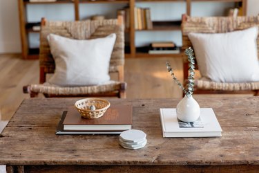 Two stacks of books on a rustic coffee table. On one, a small white vase with eucalyptus. On the other, a small woven basket. There is also a stack of grey ceramic coasters. Behind the coffee table, two chairs with light grey accent pillows.