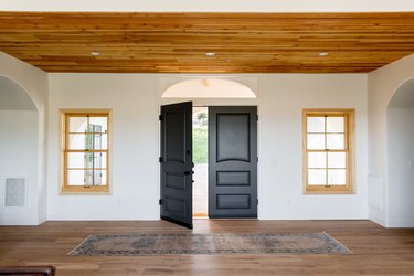 An entry hall of a Spanish-style home with double dark wood doors, one of which is open, on a white wall. Two windows with light wood frames are on either side of the door. The floor and ceiling are both made of wood. There's a rug in front of the door.