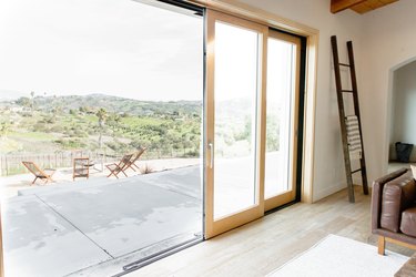 Glass sliding doors with a light wood frame are half-open, leading from a living room to an outdoor patio. The walls of the living room are white and there's a brown ladder leaning against the wall. A white and grey striped throw blanket hangs on the middle rung of the ladder. A brown leather couch is partially visible, as is a white rug. Outside, four lounge chairs are visible. Beyond them, green rolling hills.