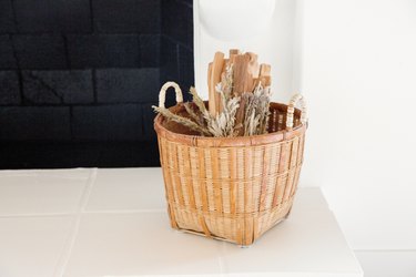 A woven basket with wood and other kindling on white floor in front of a black fireplace.