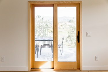 Glass sliding doors with a wood frame on a white wall. Through the glass you can see a patio with a metal table and chairs and hills in the distance.
