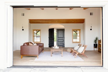 A large rectangular open doorway leading to a sitting room with a brown leather couch, a natural wood coffee table, and two wooden chairs with white pillows on a white rug. The walls of the room are white. Two windows and a dark wood double door are on the opposing wall.