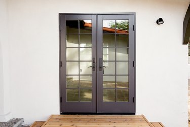 White walls with black metal double doors with glass panes, through which a brick tile courtyard is visible. Wood steps leading up tot doors.