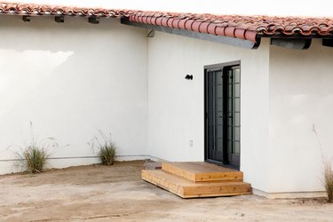 Back entrance to a white Spanish-style home with a clay tile roof. Small Mexican feather plants run along the side of the home. Two wooden steps lead up to a black sliding door.