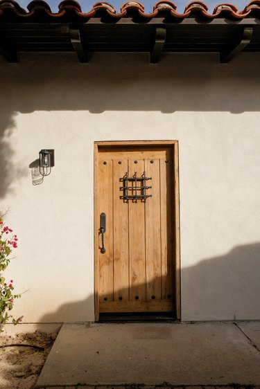 Entrance to a Spanish-style home with white walls and a clay tile roof. The door is natural wood with black metal details. There is a black sconce to the left of the door. Shadows from the roof are cast onto the wall.