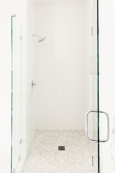 A glass shower door with a chrome handle in a white bathroom. Behind it, white shower with chrome fixtures and a white and grey patterned tile floor.
