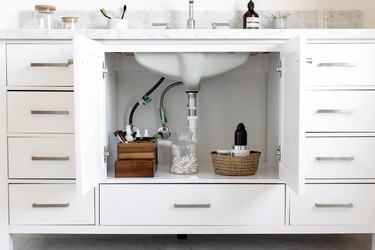 Plumbing of a sink inside a vanity cabinet. Beside are a wood makeup organizer, glass container of q-tips, a woven basket with dispensers.