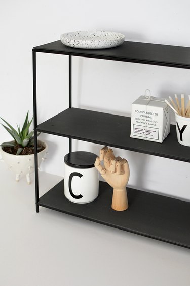 Black metal shelf with mugs and drafting tools in a white room next to small potted succulent in white plant holder