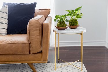 Marble side table with gold metal legs and two small potted plants beside tan leather couch in white-walled room with white rug.