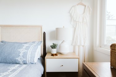 Bed with cane headboard, and blue bedding with white embroidery. Wood nightstand with a white drawer, cactus, and a white lamp.