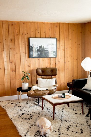 Wood paneled walls in a living room with a leather accent chair, wood Midcentury coffee table, black accents, and a dog