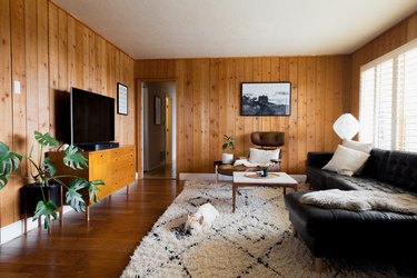rustic decor in wood paneled room with flat screen tv, natural shag rug, dog, black leather couch, plant, mid century modern lounge chair, black and white photo on wall.