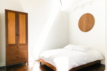 A wood armoire by a bed with white bedding. A wood circular wall hanging is above.