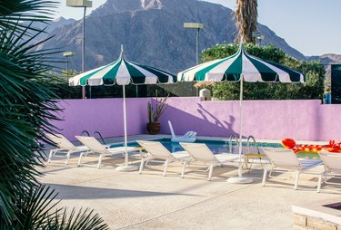 Outside shot of swimming pool, pool chairs, patio umbrellas, and pink-painted brick walls against mountains, trees, and light clouds