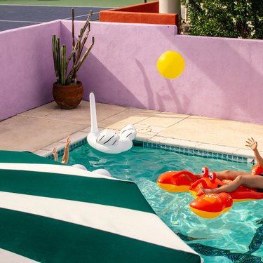 Exterior swimming pool with lobster and swan inflatables with green and white umbrella, pink-painted brick wall, and yellow balloon