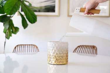 Hand pouring water from a white pitcher into a textured gold-white vase