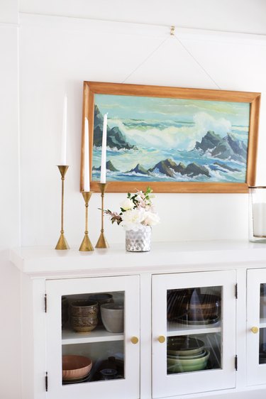 White dishware cabinet with gold candlesticks, vase of flowers, and seascape painting