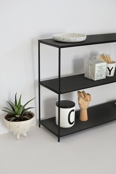 Black metal shelf with mugs and drafting tools in a white room next to small potted succulent in white plant holder