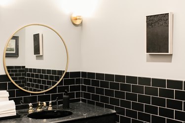 Round gold frame mirror over a black sink counter, walls with black subway tiles, a black painting.