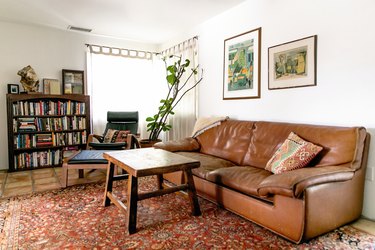 Living room with brown leather sofa, accent pillows, wood coffee table, floral rug, wood bookcase, green accent chair, art and houseplant