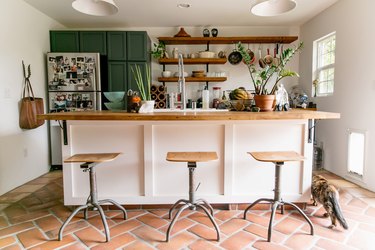 Kitchen with terra-cotta floor, white kitchen island, industrial wood stools, a white bell pendant light, green cabinets and wood shelves.