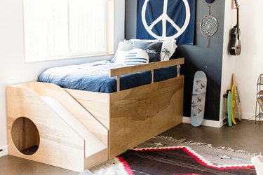 Kid's wood bed frame, with a slide, blue bedding, eclectic pillows. Peace sign flag, dream catcher, Southwest rug, and skateboard with cats.