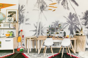Kid's playroom with palm tree wallpaper, wood table, modernist chairs, toy cars, play kitchen, wood fish mobile, and colorful Southwest rug.