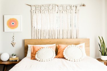 A bed with a wood headboard and peach-white pillows. A macrame curtain behind, and plants on wood night tables. Colorful wall art.