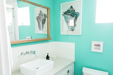 A bathroom with a turquoise wall, white tile backsplash, and white vanity. A seashell artwork and a wood frame mirror.
