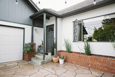 A gray and white house with a picture window and brick garden boxes. An organic paving tile and cacti in planters.