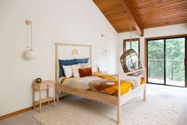 Bedroom with wood ceiling, casement windows. Neutral bedding, fiber rug, globe sconce lights, hanging boho chair.