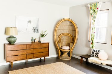 Corner of bedroom with wood mid-century sideboard, rattan throne chair, green table lamp, and 70s botanical curtains