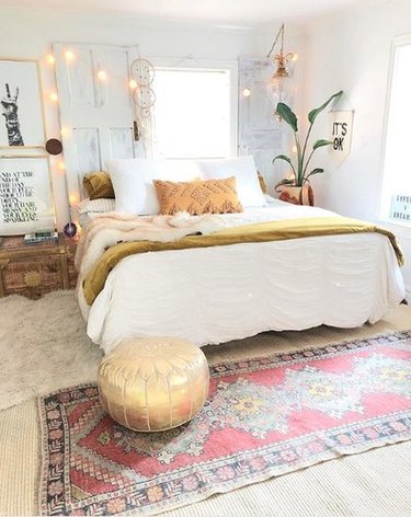 Bohemian bedroom with multiple rugs, white globe string lights, plants, and gold pouf