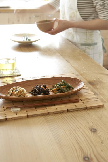 Japanese style vegetables in wooden tray with chopsticks on wooden table, woman preparing food in background