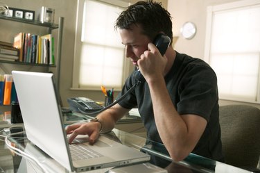a young caucasian man sits working at his computer and on the phone in his home office