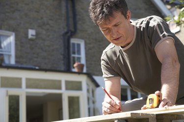 Man leaning on workbench in garden, using pencil and tape measure