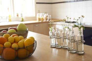 lemons and oranges in wire basket, apples in wooden bowl and flowers in glass bottle vases on wooden counter in kitchen