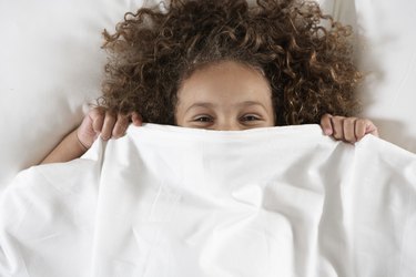 Girl (5-7) covering face with sheet in bed, portrait, overhead view