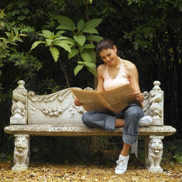 Front view of young woman sitting on bench, reading newspaper