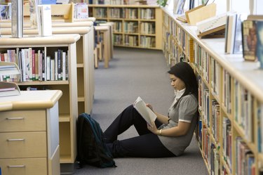 School girl (14-15) reading on floor by bookshelf in library