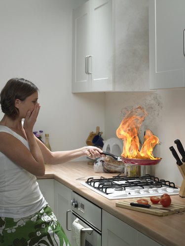 "Woman with hand to face holding burning frying pan in domestic kitchen, side view"