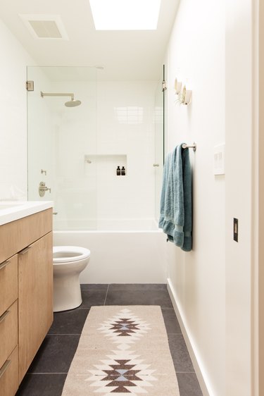 bath/shower combination with glass door, silver overhead shower head, silver shower faucet and handle, white toilet, wood vanity, gray square floor tiles, runner rug with Aztec pattern