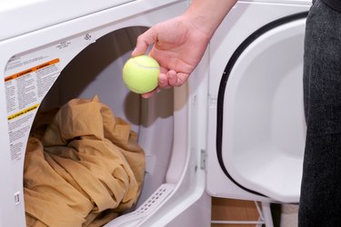 A woman's hand tossing in a green tennis ball into the dryer along with a set of sheets