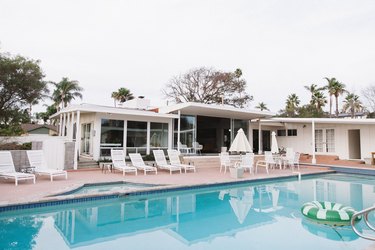 A white mid-century modern home with an in-ground swimming pool and a patio that has several white patio chairs