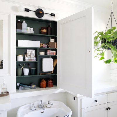 white bathroom, white subway tile, window with black trim, hanging plant, recessed medicine cabinet with painted wood green interior, white pedestal sink