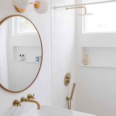 white ceramic vessel sink, gold faucet, round mirror with gold trim, white and round light fixture, white tiled shower with gold fixtures