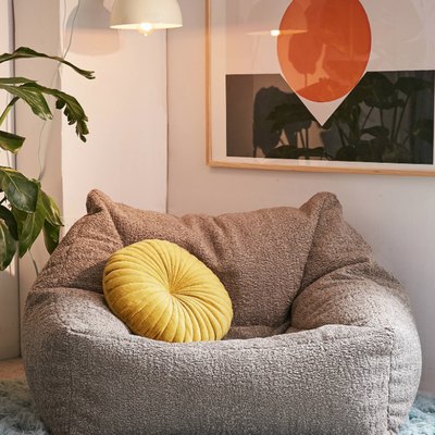 Grey faux sheepskin lounge chair, yellow pillow, light blue shag rub, white pendant lamp, plant, art with orange circle and black band.