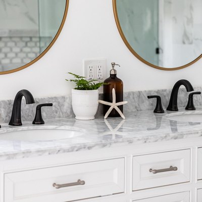 white marble countertop, two recessed sinks with black hardware, two round mirrors with gold trim